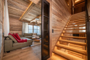 Alpines Wohnzimmer mit massivem, dynamisch geformtem Salontisch aus Kernesche, rotem Wollfilzkissen und grünem Ledersofa, Eichen­treppe mit Stahlgeländer, Eichendielen und Lofttüren im Stahllook.