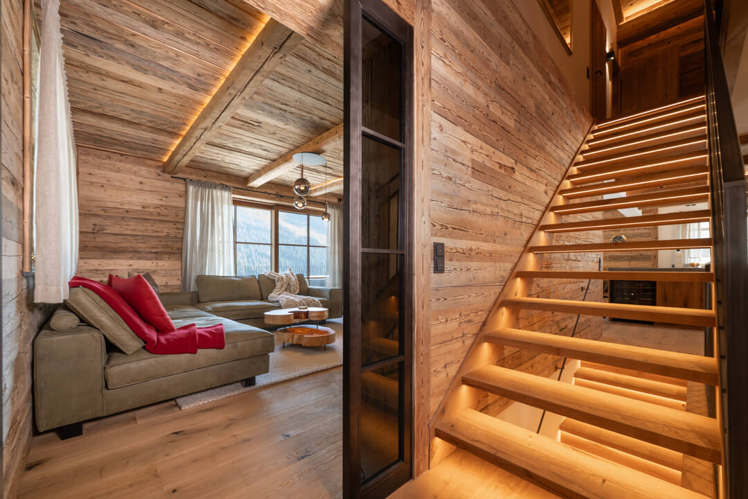 Alpines Wohnzimmer mit massivem, dynamisch geformtem Salontisch aus Kernesche, rotem Wollfilzkissen und grünem Ledersofa, Eichen­treppe mit Stahlgeländer, Eichendielen und Lofttüren im Stahllook.