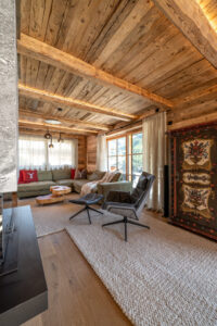 Alpines Wohnzimmer mit massivem, dynamisch geformtem Salontisch aus Kernesche, Weißer Wollfilzteppich, grünem Ledersofa und Lederfauteuil. Beleuchtete Altholzdecke setzt warme Akzente.