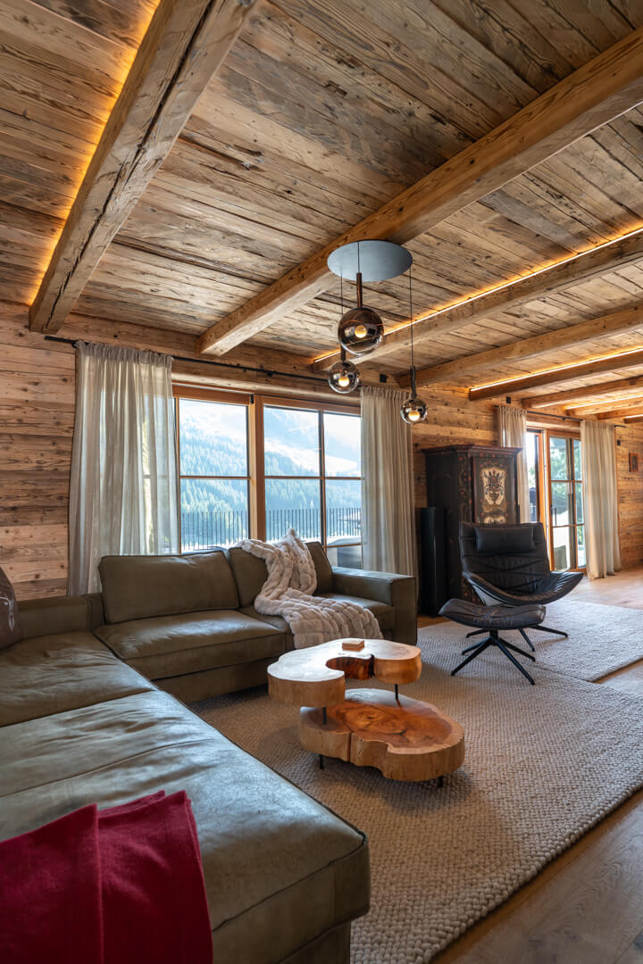 Alpines Wohnzimmer mit massivem, dynamisch geformtem Salontisch aus Kernesche, grünem Ledersofa und Lederfauteuil. Beleuchtete Altholzdecke