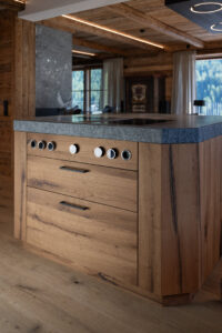 Chaletküche mit Kücheninsel aus massiver Eiche, rauer Arbeitsplatte aus Rauriser Naturstein und modernem Bora-Kochfeld.