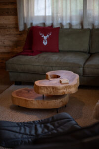 Alpines Wohnzimmer mit massivem, dynamisch geformtem Salontisch aus Kernesche, rotem Wollfilzkissen mit Hirschmotiv und grünem Ledersofa.
