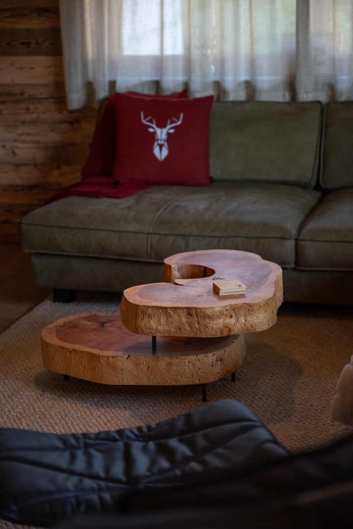 Alpines Wohnzimmer mit massivem, dynamisch geformtem Salontisch aus Kernesche, rotem Wollfilzkissen mit Hirschmotiv und grünem Ledersofa.