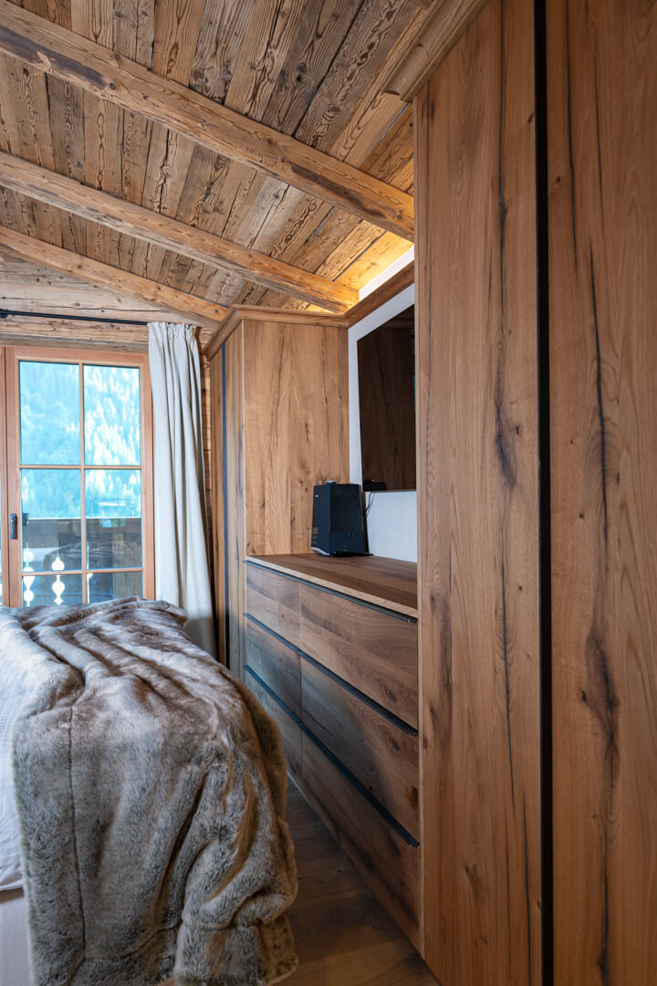 Einbauschränke im Einbauschränke im alpinen Schlafzimmer aus Eiche mit subtilen Stahlgriffen, gemütlicher Felldecke, hellen Gardinen und Altholzverkleidung.