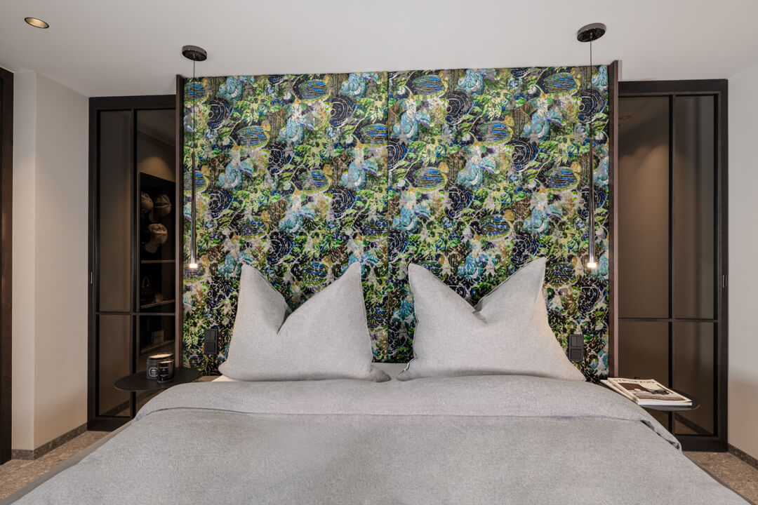 Schlafzimmer mit einem gepolsterten Headboard mit floralem Print; dahinter ein begehbarer Kleiderschrank mit dunkelbraunen Glastüren.“