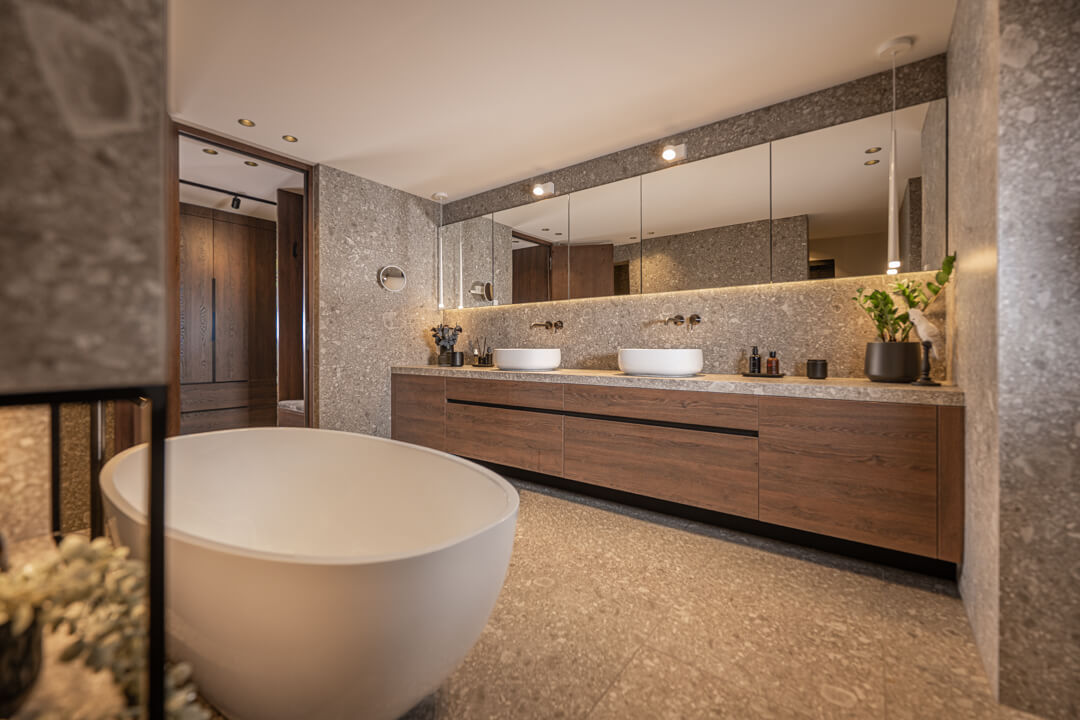 Badezimmer mit marmorierten Naturstein-Look-Fliesen in Ivory, dunkelbraunem Holz-Waschtisch, freistehender Badewanne und weißen Akzenten in Hängelampen.