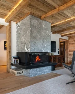 Massiver Kamin aus Rauriser Naturstein im Chalet-Wohnzimmer mit alpinem Interieur, Altholzdecke und weißem Wollteppich.