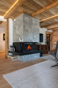 Massiver Kamin aus Rauriser Naturstein im Chalet-Wohnzimmer mit alpinem Interieur, Altholzdecke und weißem Wollteppich.